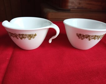 Puede incluir: Dos tazas de cerámica blanca con un patrón floral dorado. Las tazas están sobre una superficie roja.