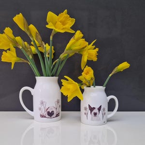 May include: Two white ceramic vases with floral designs. The larger vase is filled with yellow daffodils and the smaller vase has a single yellow daffodil. Both vases feature a dog illustration.