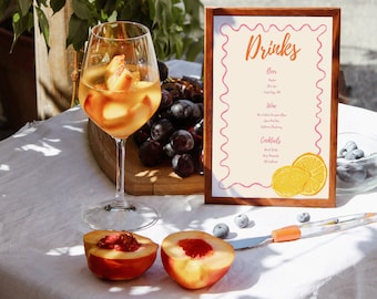 May include: A refreshing drink with peach slices in a wine glass, next to a wooden-framed menu board with the word "Drinks" and a list of beverages. Halved peaches and grapes are also on the table, along with a knife and blueberries.