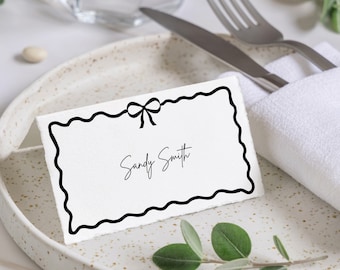May include: A white place card with a black wavy border and a black bow at the top. The text "Sandy Smith" is written in black ink on the card.