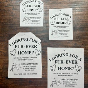 May include: Four white house-shaped signs with black text and graphics. Each sign says "Looking for a Fur-Ever Home?" and includes the text "Let me help with you all your real estate needs!"  The signs also include the name "Megan Smithson", the text "The Realty Group", an email address, and a phone number.  The signs also include a graphic of a key and a house.