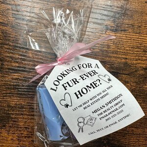 May include: A white card with black text that reads "Looking for a fur-ever home? Let me help with all your real estate needs! Megan Smithson The Realty Group email@email.com 205-555-5555 Call, text, or email anytime!" The card is tied with a pink ribbon to a blue plastic bag with a dog poop bag dispenser.
