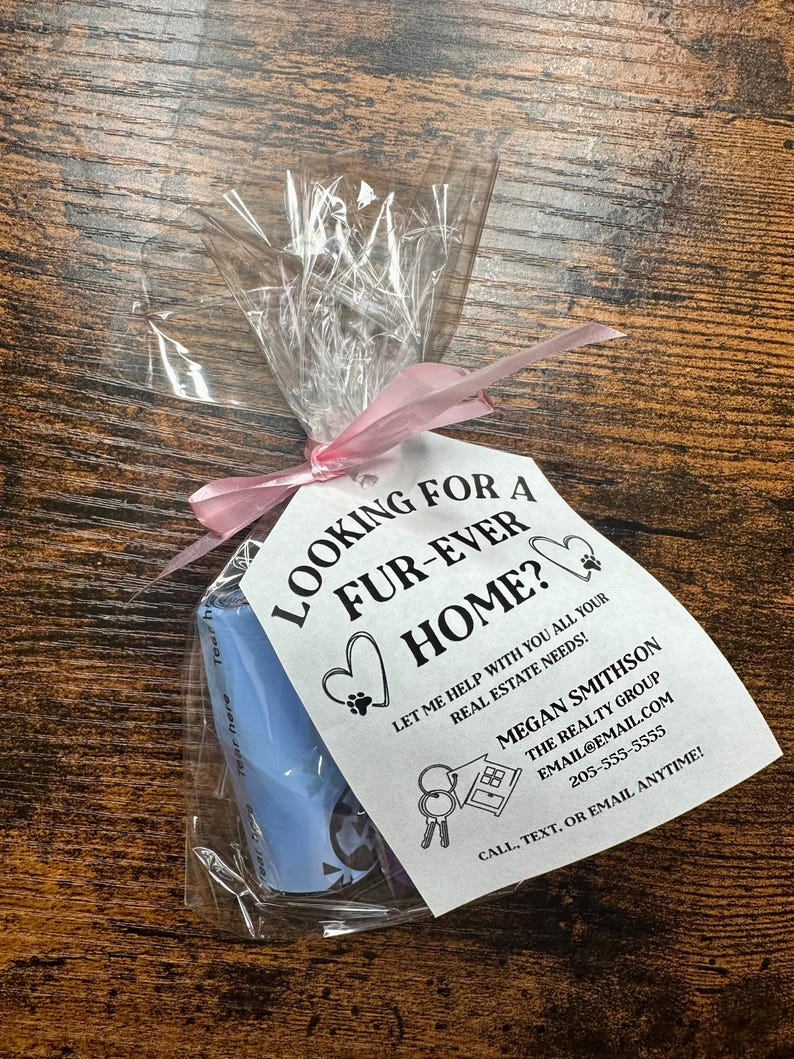 May include: A white card with black text that reads "Looking for a fur-ever home? Let me help with all your real estate needs! Megan Smithson The Realty Group email@email.com 205-555-5555 Call, text, or email anytime!" The card is tied with a pink ribbon to a blue plastic bag with a dog poop bag dispenser.