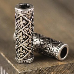May include: Two silver-colored cylindrical beads with a fleur-de-lis pattern. The beads are resting on a wooden surface. The design features a repeating diamond pattern with the fleur-de-lis symbol inside each diamond. The beads have a detailed, antique look.