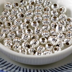 May include: A white bowl filled with silver metal beads. The beads have a ribbed texture and are round in shape.
