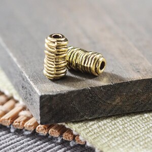 May include: Two gold-colored metal beads with a textured surface. The beads are on a dark brown wooden surface.