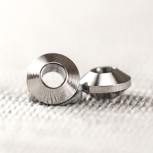 May include: Two small, shiny, silver metal washers with a beveled edge and a central hole. The washers are on a textured, off-white surface. These washers are likely used in construction or mechanical applications.