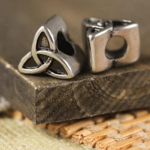 Può includere: Due perline color argento. Una perlina presenta un design a nodo celtico, simbolo di interconnessione. L'altra perlina è di forma quadrata con un foro circolare. Le perline sono esposte su una superficie di legno.