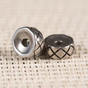 May include: Two silver-colored metal beads with a diamond pattern. One bead has a flared opening, while the other is flat. The beads are likely for jewelry making or crafts.