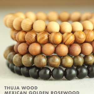 May include: Five wooden bead bracelets stacked on top of each other. The bracelets are made of different types of wood, including thuja wood, Mexican golden rosewood, brown sandalwood, green sandalwood, and Indonesian ebony sandalwood.