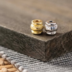 May include: Two metal beads, one gold and one silver, with a decorative pattern. The beads are on a wooden surface.