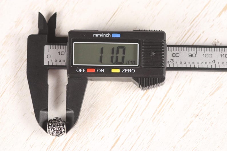 Op de afbeelding: Een digitale schuifmaat meet een kleine, zilveren kraal met een schedelontwerp. Het digitale display toont 11,0 mm. De schuifmaat is zwart, de achtergrond is licht hout.