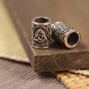 Può includere: Due perline in metallo argentato con intricati disegni di nodi celtici. Le perline sono cilindriche e hanno una finitura liscia e lucida.