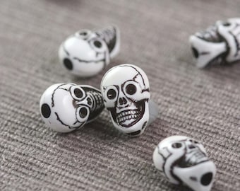 May include: A collection of white skull-shaped beads with black painted details. The beads are scattered on a grey textured surface.
