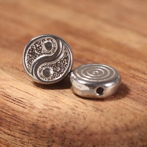 May include: Two silver-colored beads. One bead features a Yin Yang symbol, the other has a spiral design. The beads are round and have a small hole for stringing. They are displayed on a wooden surface.