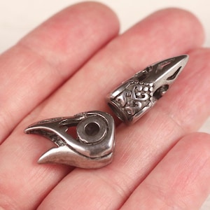 Puede incluir: Dos cuentas plateadas con forma de cráneo de cuervo. Una cuenta es un cráneo completo, mientras que la otra es un cráneo parcial. Las cuentas tienen detalles intrincados y probablemente son para hacer joyas o manualidades.