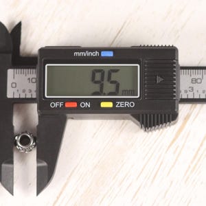 Op de afbeelding: Een zwarte digitale schuifmaat meet een kleine, zilverkleurige kraal. De schuifmaat geeft een meting van 9,5 mm weer. De schuifmaat staat op een lichtgekleurd houten oppervlak.