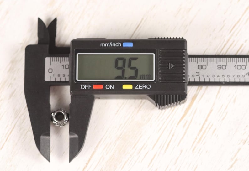 Op de afbeelding: Een zwarte digitale schuifmaat meet een kleine, zilverkleurige kraal. De schuifmaat geeft een meting van 9,5 mm weer. De schuifmaat staat op een lichtgekleurd houten oppervlak.