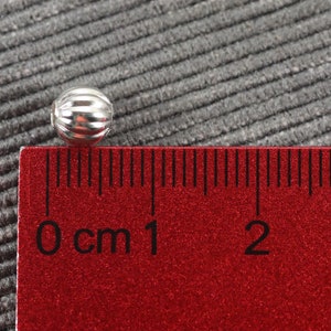 May include: A silver bead with a ribbed texture, measuring approximately 1 cm in diameter, is placed on a red ruler with black markings.