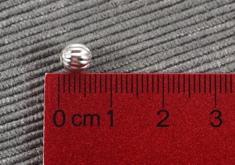 May include: A silver bead with a ribbed texture, measuring approximately 1 cm in diameter, is placed on a red ruler with black markings.