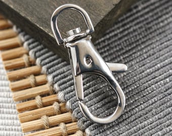 May include: A silver metal trigger snap hook with a rounded opening. The hook is lying on a woven surface.