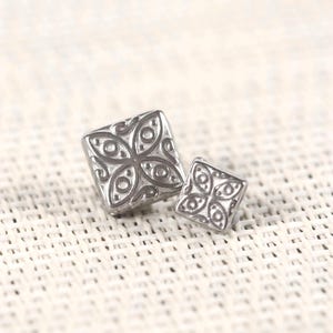 May include: Two square, silver-colored buttons with an embossed floral design. The buttons are different sizes, with the larger one in the foreground. The background is a light, textured surface.