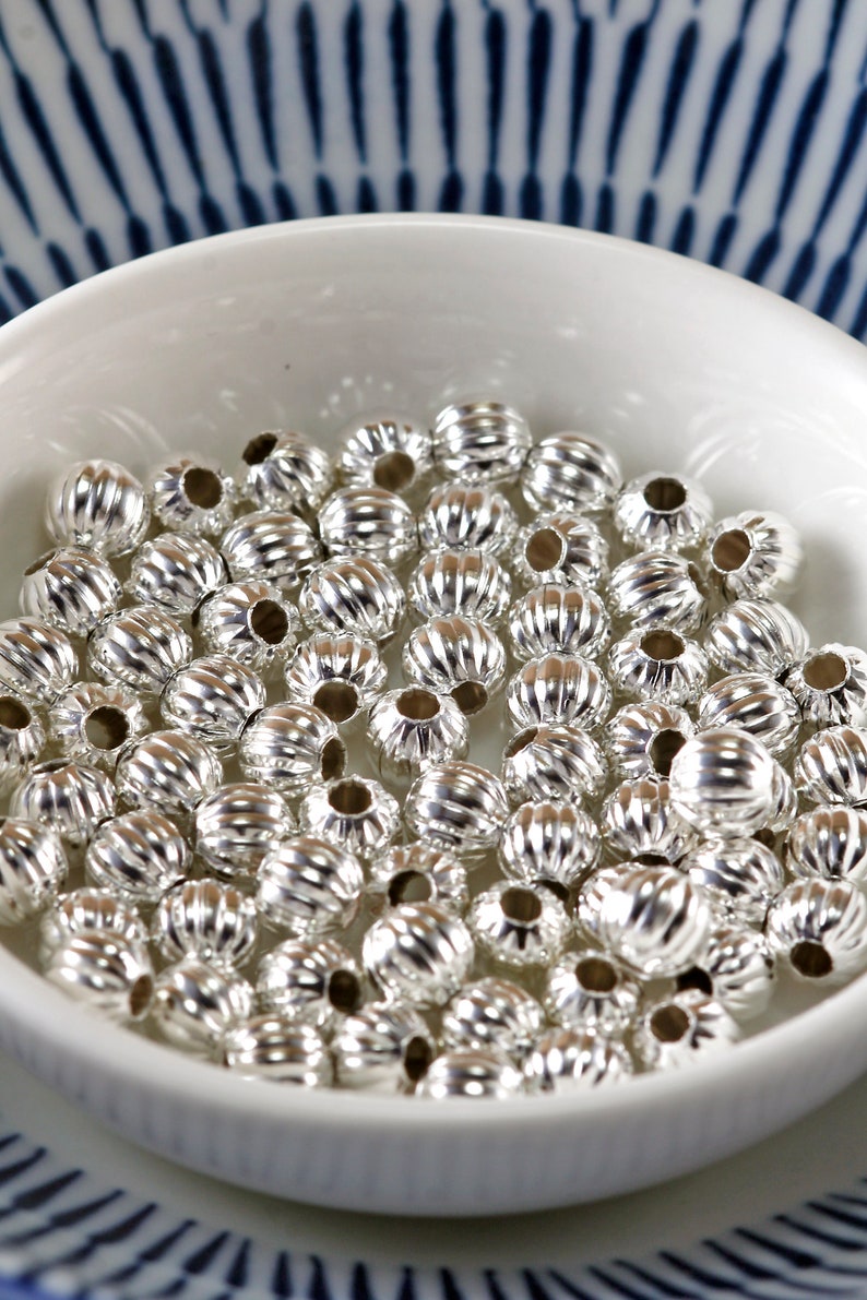 May include: A white bowl filled with silver metal beads. The beads have a ribbed texture and are round in shape.