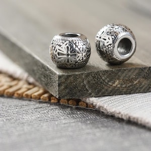 May include: Two silver-colored, round beads with intricate floral and dot patterns. The beads have a hollow core, and are displayed on a wooden surface. The beads are likely for jewelry making or crafting.