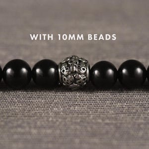 Op de afbeelding: Een close-up van een zwarte kralenarmband met een decoratieve zilveren kraal in het midden. De armband heeft kralen van 10 mm en wordt gepresenteerd op een getextureerd grijs oppervlak. De tekst "WITH 10mm BEADS" staat boven de armband.