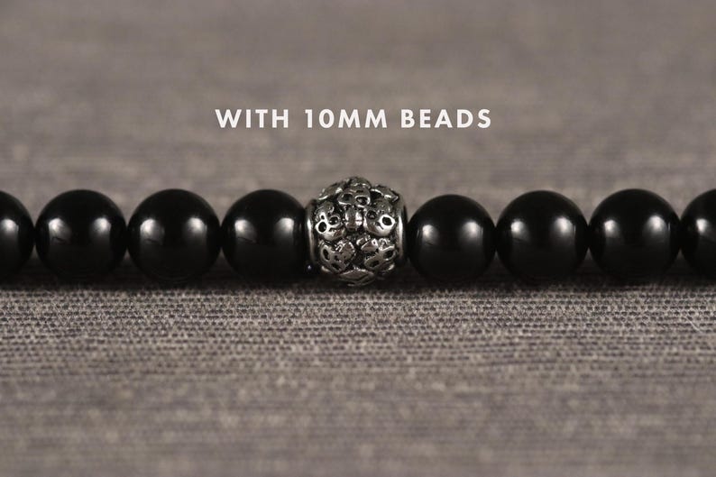 Op de afbeelding: Een close-up van een zwarte kralenarmband met een decoratieve zilveren kraal in het midden. De armband heeft kralen van 10 mm en wordt gepresenteerd op een getextureerd grijs oppervlak. De tekst "WITH 10mm BEADS" staat boven de armband.