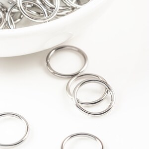 May include: A white bowl filled with silver jump rings. Several jump rings are scattered on a white surface.