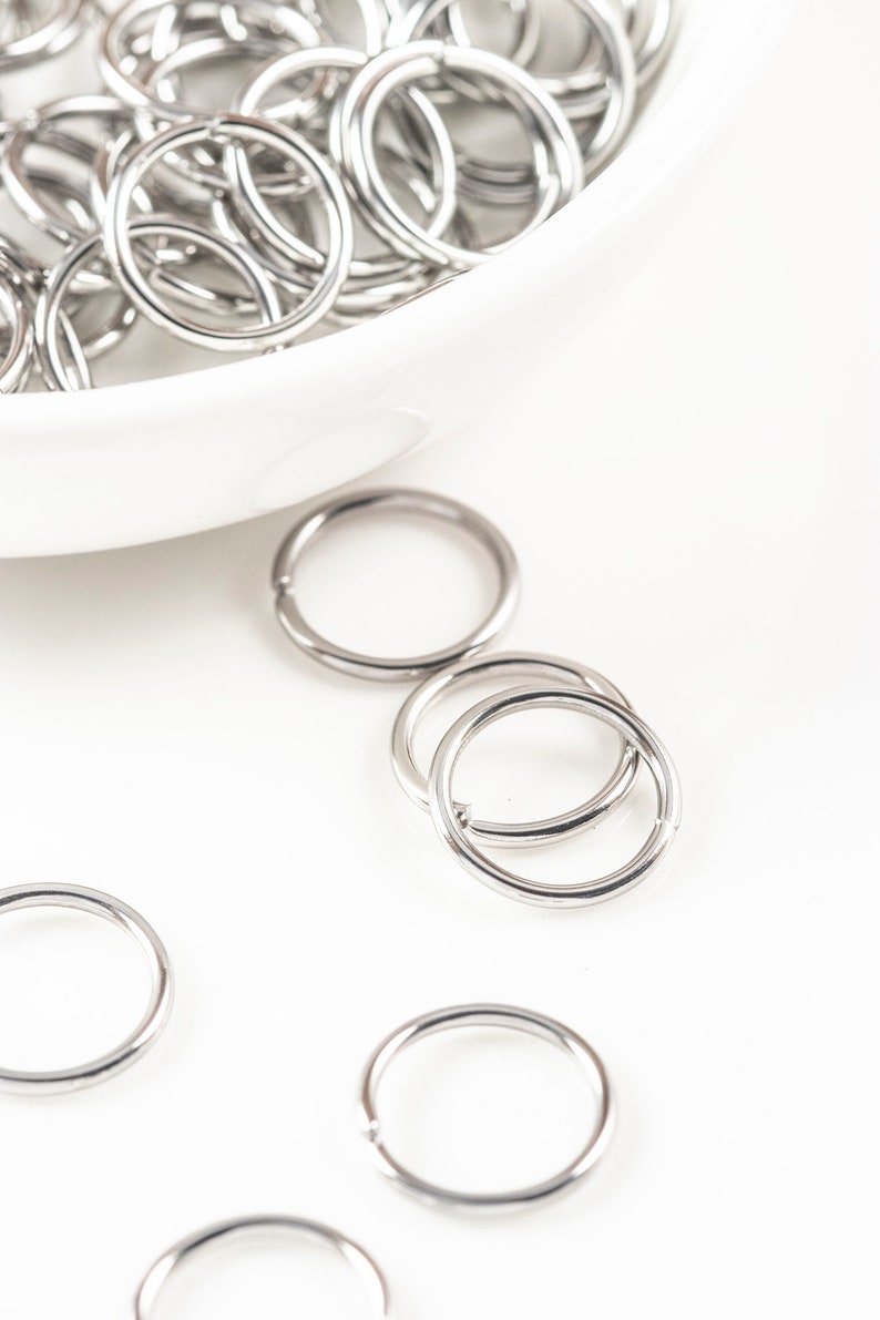 May include: A white bowl filled with silver jump rings. Several jump rings are scattered on a white surface.