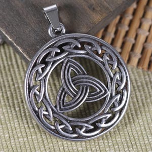 Può includere: Ciondolo con nodo celtico color argento. Il ciondolo presenta un design circolare con un intricato motivo a nodo intrecciato, incluso un simbolo centrale della triquetra. Ha un piccolo anello per una catena e viene presentato su una superficie di legno.