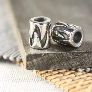 May include: Two silver-colored cylindrical beads with a repeating flame-like pattern. The beads are lying on a dark gray, rectangular surface. The beads have a hole running through the center.