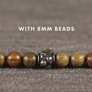 Op de afbeelding: Een close-up van een armband met 8 mm houten kralen. De kralen zijn in verschillende tinten bruin en groen, met een zilverkleurige accentkraal in het midden. De tekst "WITH 8MM BEADS" wordt boven de armband weergegeven.