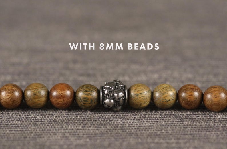Op de afbeelding: Een close-up van een armband met 8 mm houten kralen. De kralen zijn in verschillende tinten bruin en groen, met een zilverkleurige accentkraal in het midden. De tekst "WITH 8MM BEADS" wordt boven de armband weergegeven.