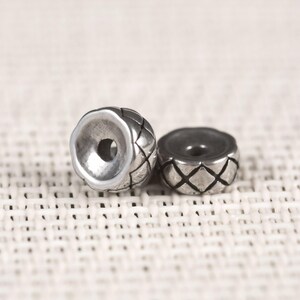 May include: Two silver-colored metal beads. One bead is upright, showing a large hole, and the other is lying on its side. Both beads have a diamond pattern etched into their surfaces. The beads are likely for jewelry making.