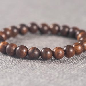 Puede incluir: Un brazalete de cuentas de madera marrón con un diseño sencillo. El brazalete está hecho de cuentas redondas que están enhebradas en un cordón.