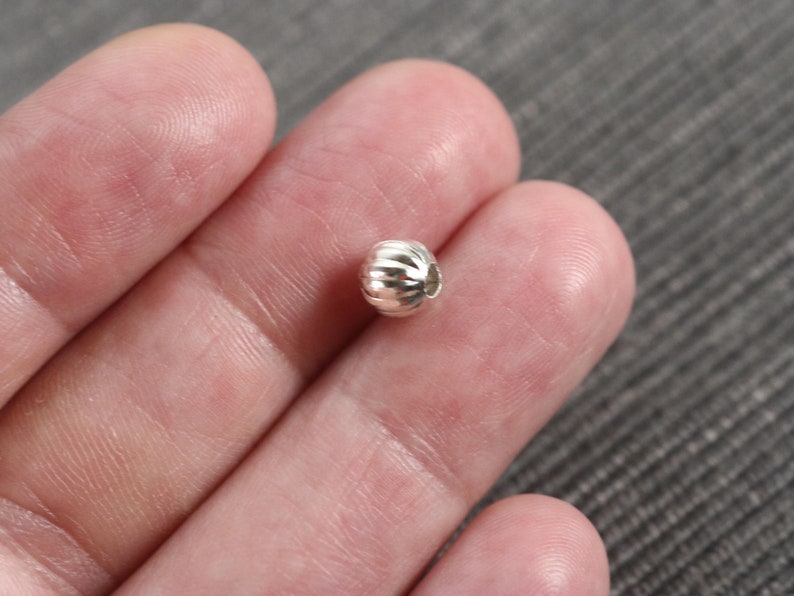 May include: A silver bead with a textured, ribbed surface. The bead is round and has a small hole for stringing.