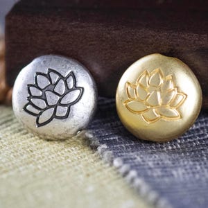 May include: Two round metal buttons with a lotus flower design. One button is silver with a black lotus flower and the other button is gold with a gold lotus flower.