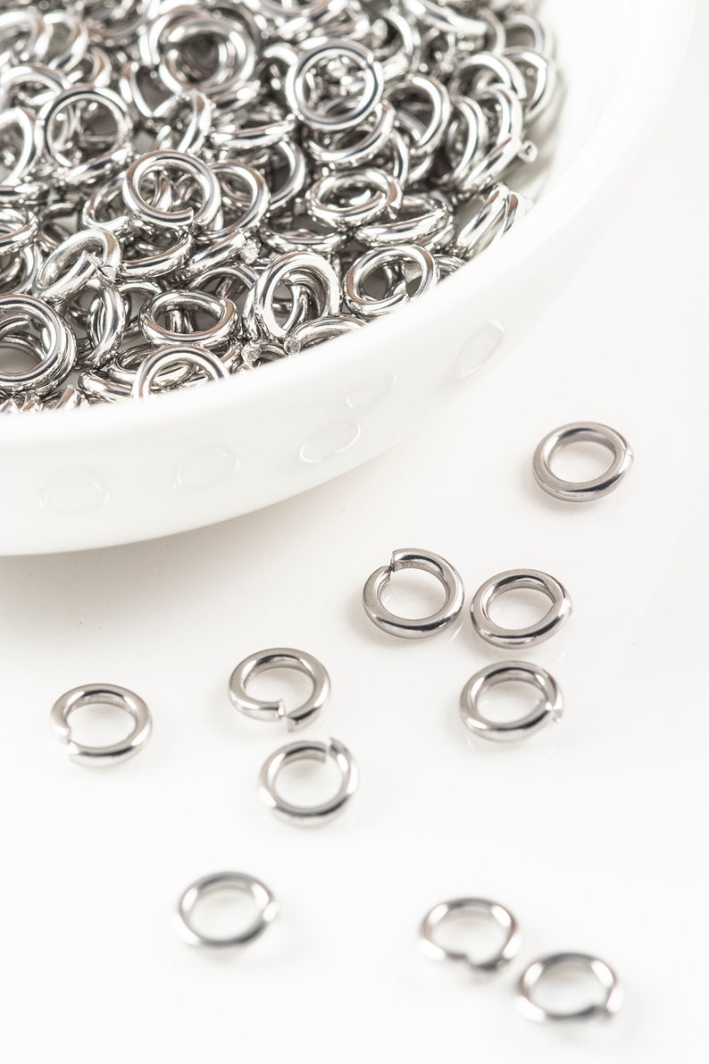 May include: A white bowl filled with silver jump rings, with additional jump rings scattered on a white surface.