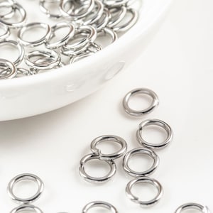 May include: A white bowl filled with silver jump rings, with more jump rings scattered on a white surface.