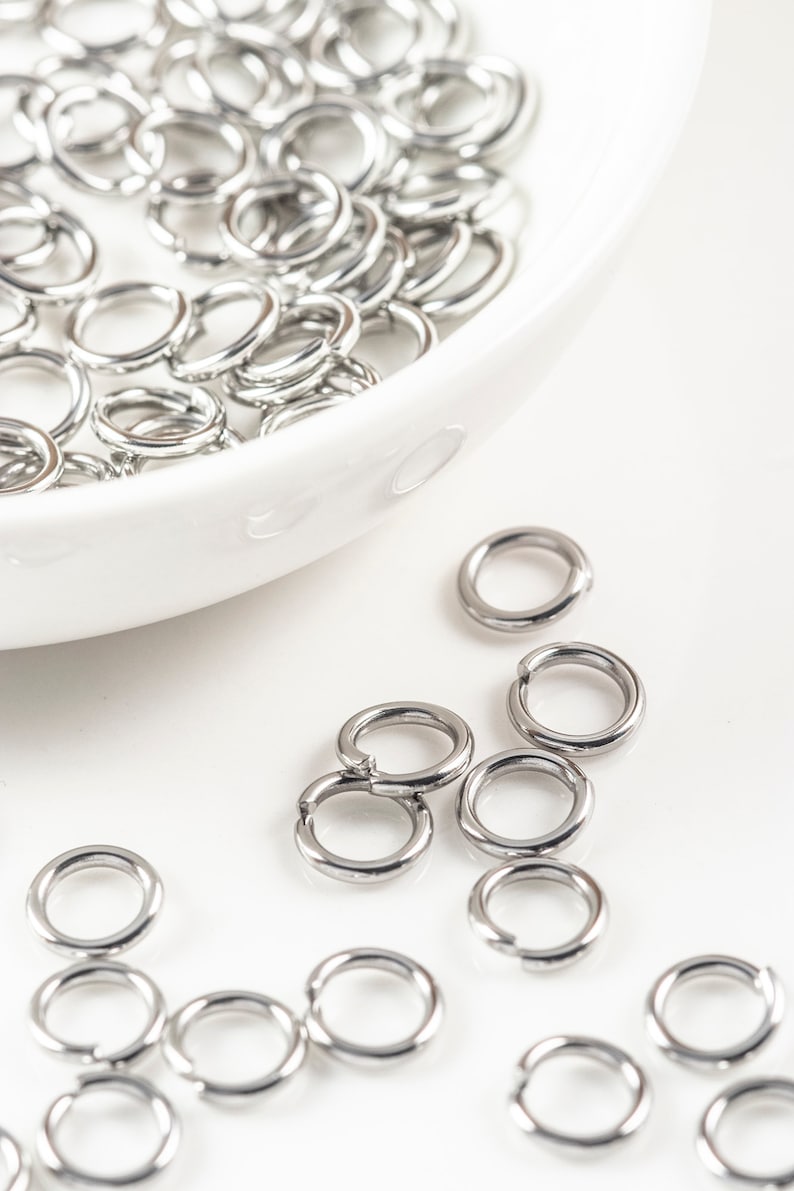 May include: A white bowl filled with silver jump rings, with more jump rings scattered on a white surface.