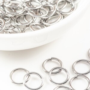 May include: A white bowl filled with silver jump rings. Several jump rings are scattered on a white surface.