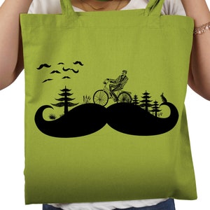 Könnte beinhalten: Eine grüne Einkaufstasche mit einer schwarzen Silhouette eines Mannes, der mit dem Fahrrad durch einen Wald fährt, mit einem großen Schnurrbart im Vordergrund. Der Mann trägt einen Hut und ein kariertes Hemd. Über dem Wald befinden sich kleine Schnurrbärte am Himmel.