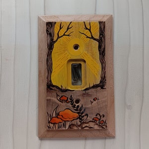 May include: A wooden light switch cover with a hand-painted design of a sun, trees, and mushrooms. The sun is yellow, the trees are black, and the mushrooms are orange and brown.