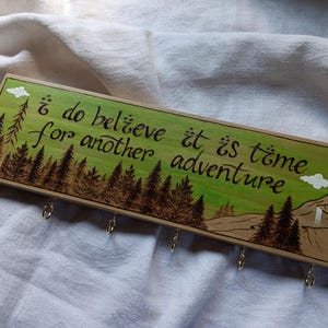 May include: Wooden key holder with a green and brown woodburned design. The design features a forest scene with trees and a mountain. The text "I do believe it is time for another adventure" is written in cursive on the wood. There are five metal hooks on the bottom of the key holder.