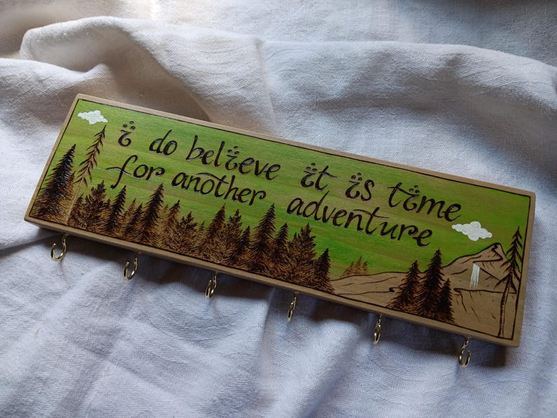 May include: Wooden key holder with a green and brown woodburned design. The design features a forest scene with trees and a mountain. The text "I do believe it is time for another adventure" is written in cursive on the wood. There are five metal hooks on the bottom of the key holder.