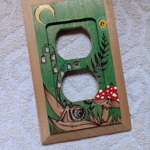 May include: Wooden electrical outlet cover hand-painted with a whimsical forest scene. The green background features a crescent moon, mushrooms, and a snail. The cover has a natural wood frame and two outlet holes.