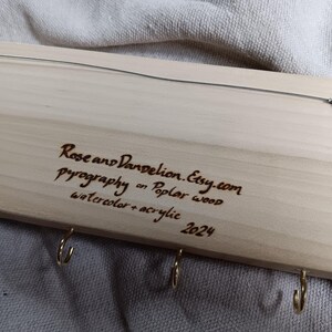 May include: Wooden key holder with five hooks and a burned-in inscription: "Rose and Dandelion.Etsy.com Pyrography on Poplar wood Watercolor + acrylic 2024"